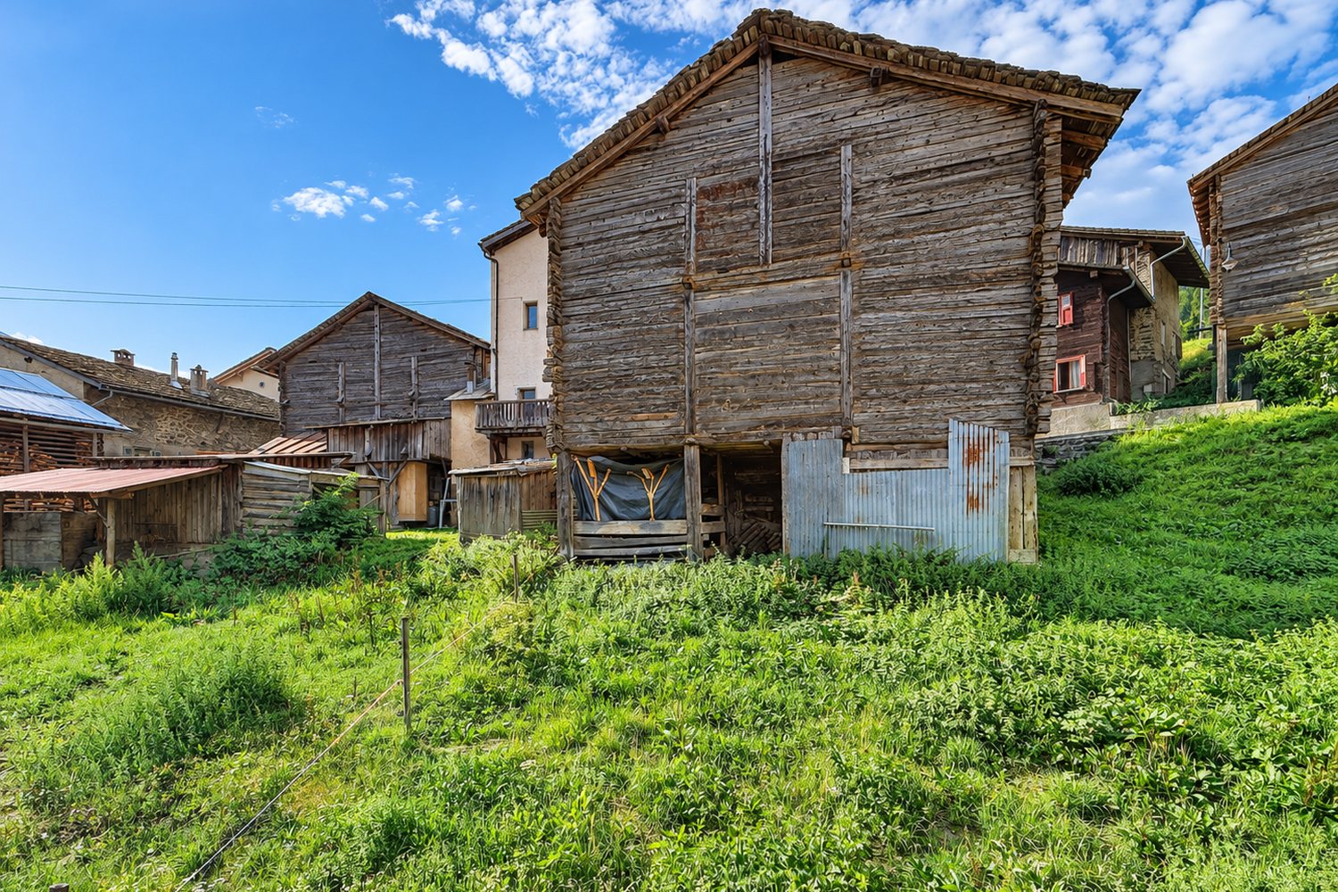 La façade aval de la grange sur ses pilotis, fermée d'éléments légers, dans l'herbage du hameau.